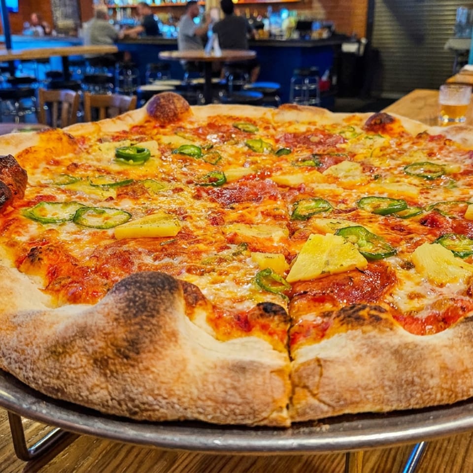 Large wood-fired pizza topped with pepperoni, pineapple, and jalapeños on a metal tray in a lively restaurant.
