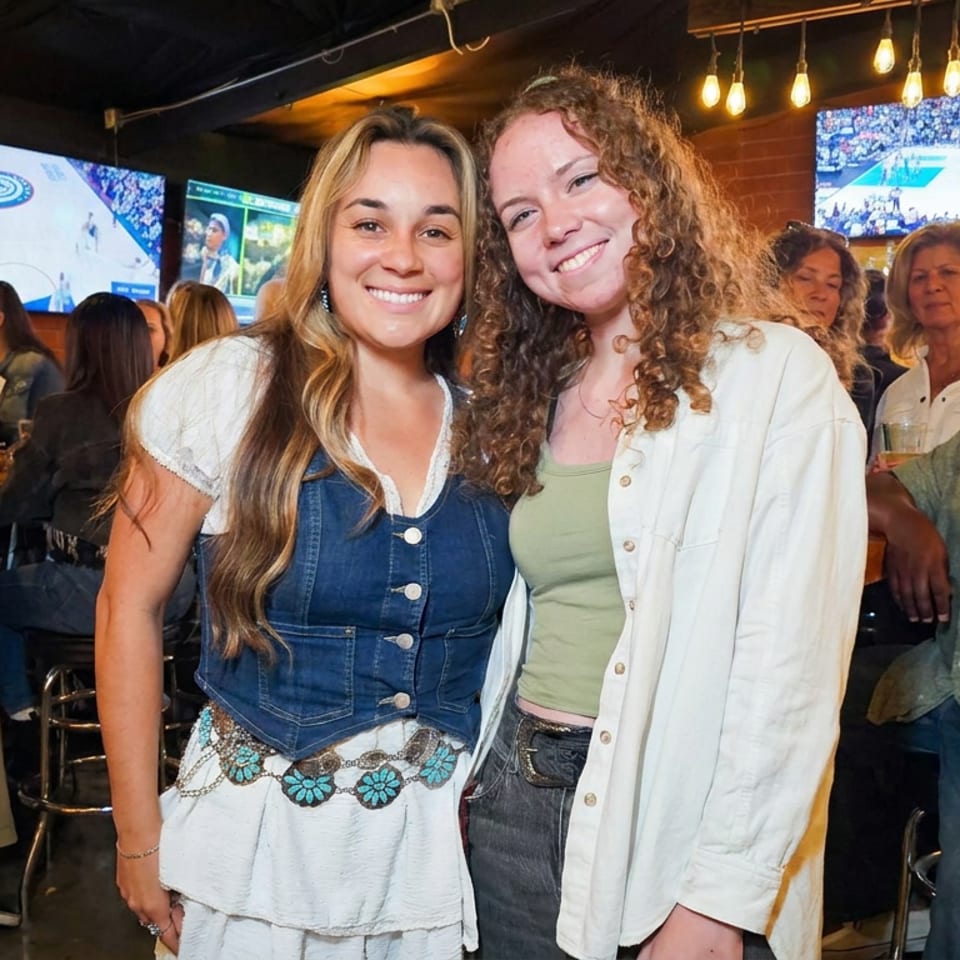Two smiling women pose together inside a busy sports bar with televisions, hanging lights, and patrons in the background.