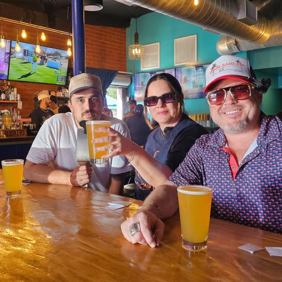 Three friends wearing sunglasses and caps raise beers at a lively bar with televisions, exposed ductwork, and warm hanging lights.
