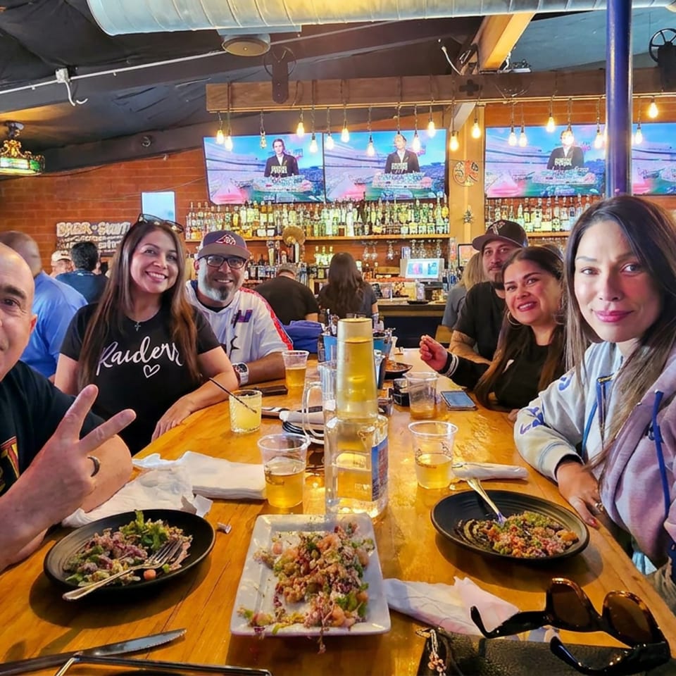 Friends pose around a restaurant table with plated food and drinks, surrounded by a lively bar, televisions, and exposed brick walls.