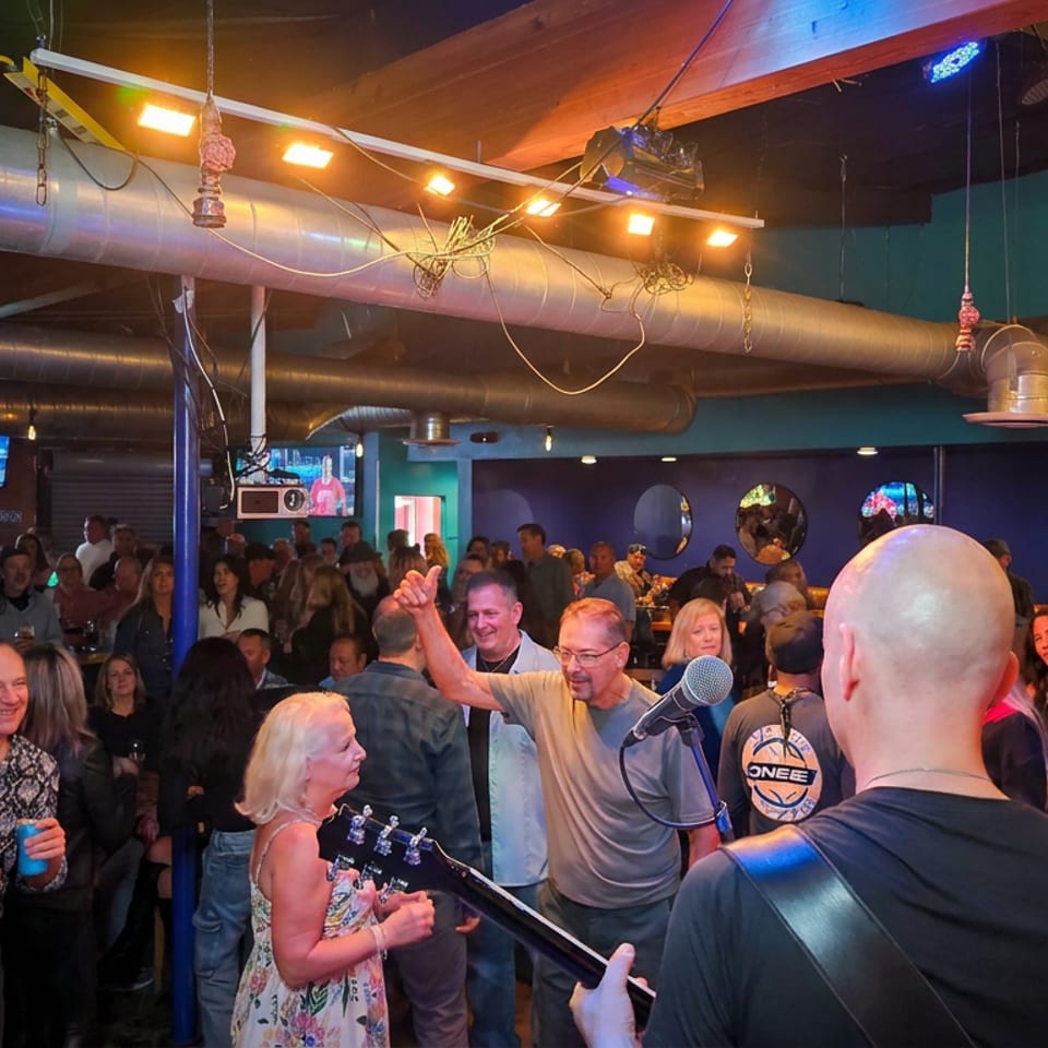 Live band performs before a lively crowd in a dimly lit restaurant bar with colorful lighting and exposed ductwork.