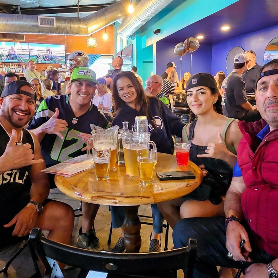Friends pose with drinks at a lively sports bar, surrounded by televisions showing a game and colorful decor.