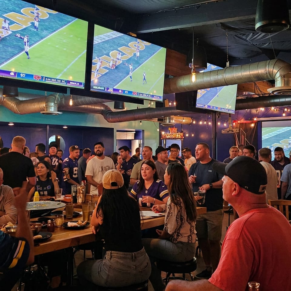 Crowded sports bar with diners watching football on multiple large screens beneath exposed ductwork and string lights.