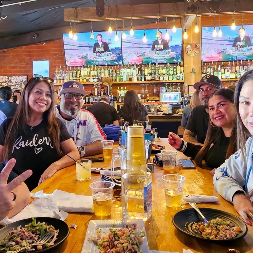 Friends gather around a restaurant table with beers and food while multiple televisions show a bartender behind the bar.