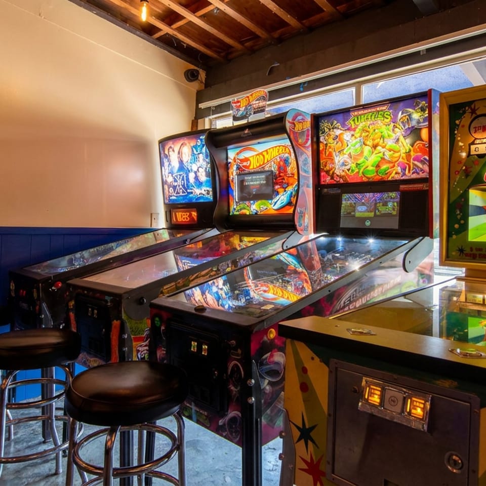 Arcade room featuring colorful pinball machines, a basketball game, and black stools beside large windows.
