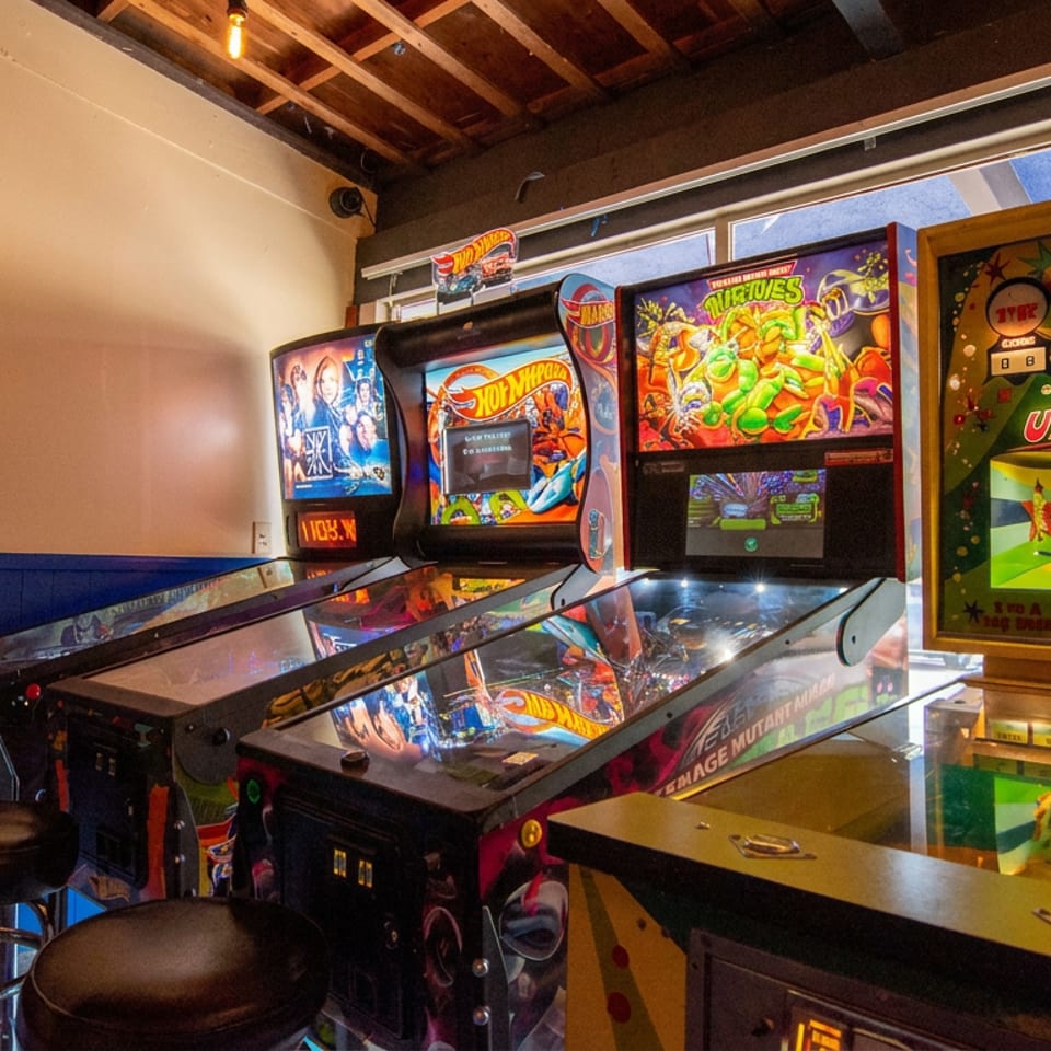 Restaurant arcade area featuring multiple colorful pinball machines, a foosball table, and bar stools beside large windows.