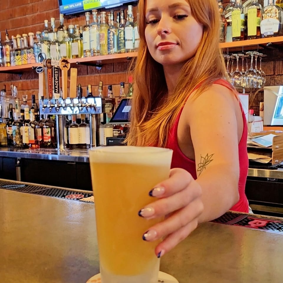 Bartender serves a tall golden beer across a polished bar, with liquor bottles, taps, and glassware lining the brick-wall backdrop.