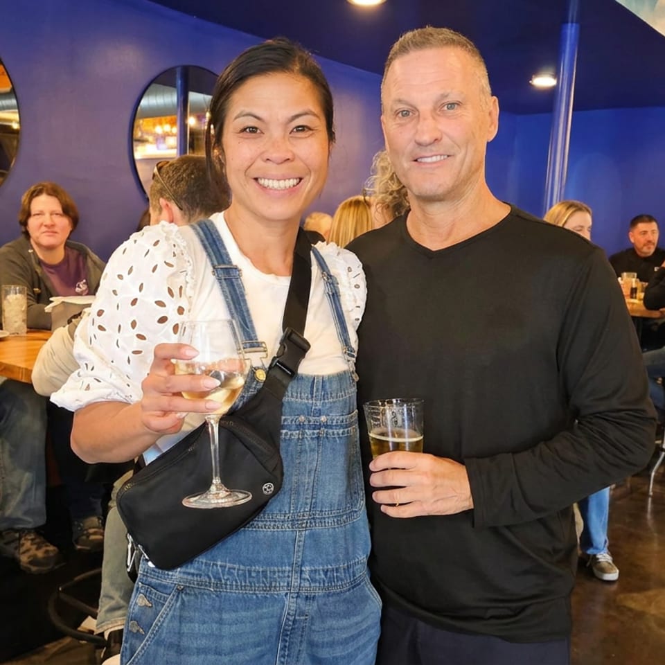 Smiling couple holding wine and beer in a lively restaurant with blue walls, seated diners, wooden tables, and bar stools.