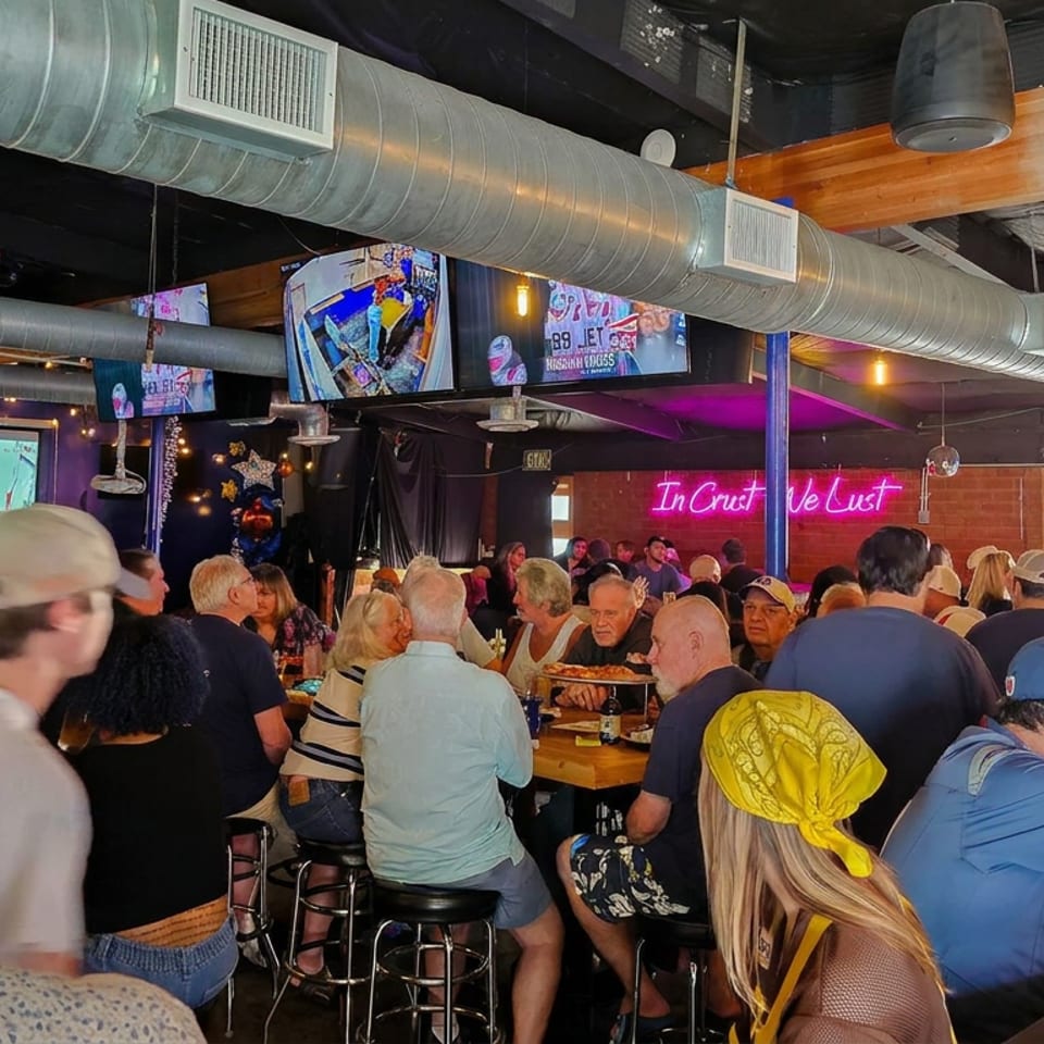 Crowded sports bar with diners, multiple televisions showing a game, exposed ductwork, neon signage, and football decorations.