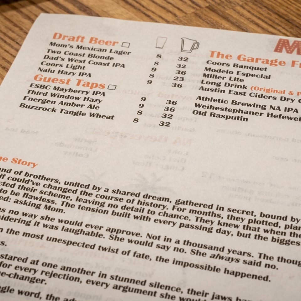 Restaurant menu on a wooden table listing draft beers, guest taps, garage fridge drinks, and the restaurant’s story.