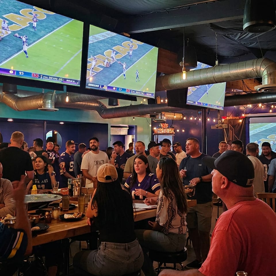 Crowded sports bar with patrons watching football on multiple large screens, surrounded by tables, drinks, exposed ductwork, and string lights.