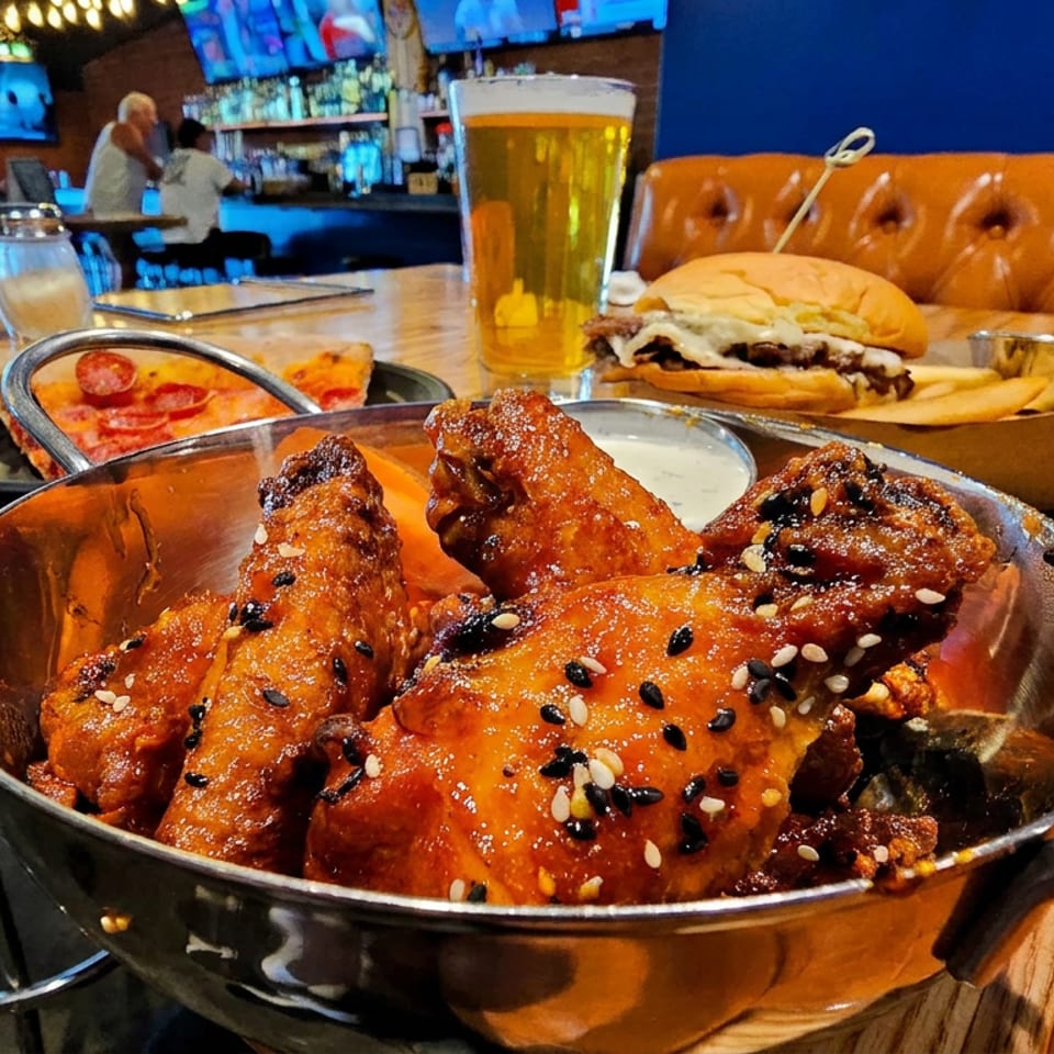 Glazed chicken wings topped with black and white sesame seeds, served alongside pizza, a burger with fries, and beer in a restaurant.