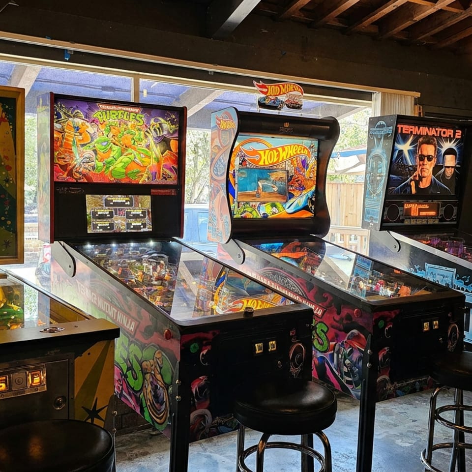 Retro arcade corner featuring Upper Deck, Turtles, Hot Wheels, and Terminator 2 pinball machines with stools by large windows.