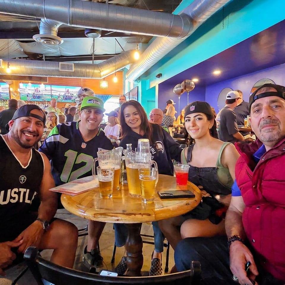 Friends pose around a table with beer in a brightly colored sports bar displaying televisions and exposed ductwork.