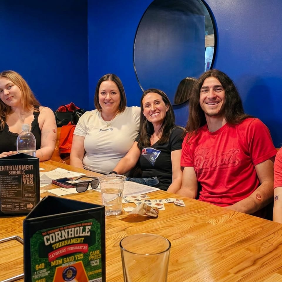 Six smiling adults pose around a wooden restaurant table with drinks, menus, and blue walls decorated with mirrors.