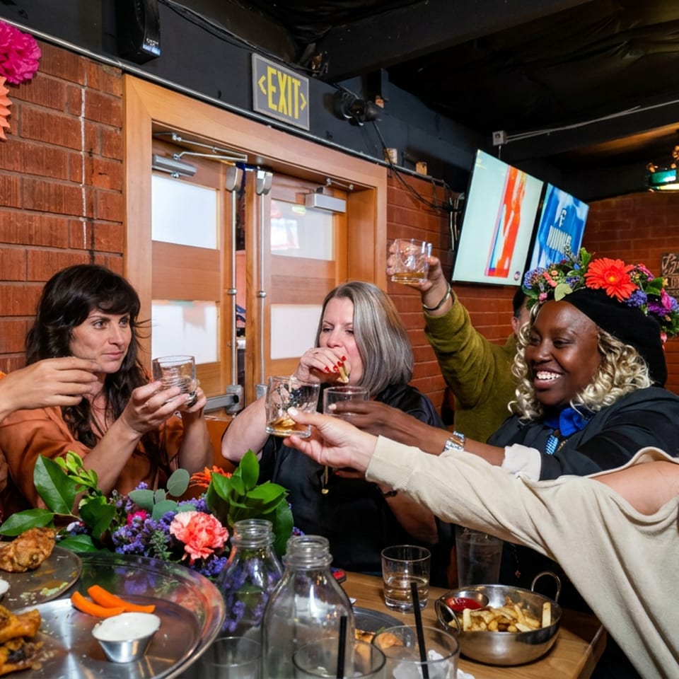 Friends toast with drinks around a restaurant table decorated with colorful flowers, appetizers, and festive wall decorations.