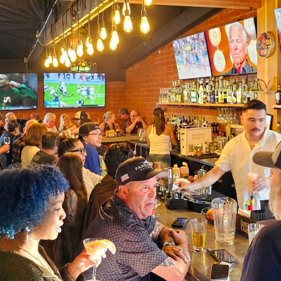 Busy sports bar with patrons gathered at the counter, bartender serving drinks, liquor shelves, warm hanging lights, and multiple televisions.