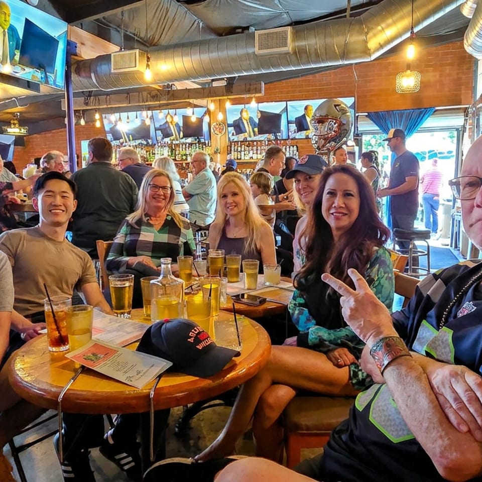 Friends gather around a table with drinks inside a lively sports bar featuring televisions, exposed ducts, and a colorful interior.